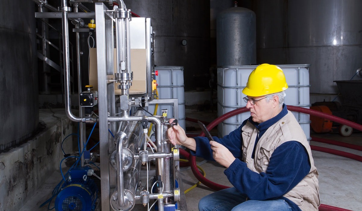 An industrial worker wearing a yellow hard hat checking gauges inside of a manufacturing plant.